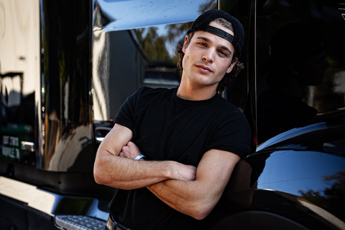 Singer-songwriter Max McNown in a backward cap and black t-shirt posing with crossed arms against a black truck for a press portrait photo.